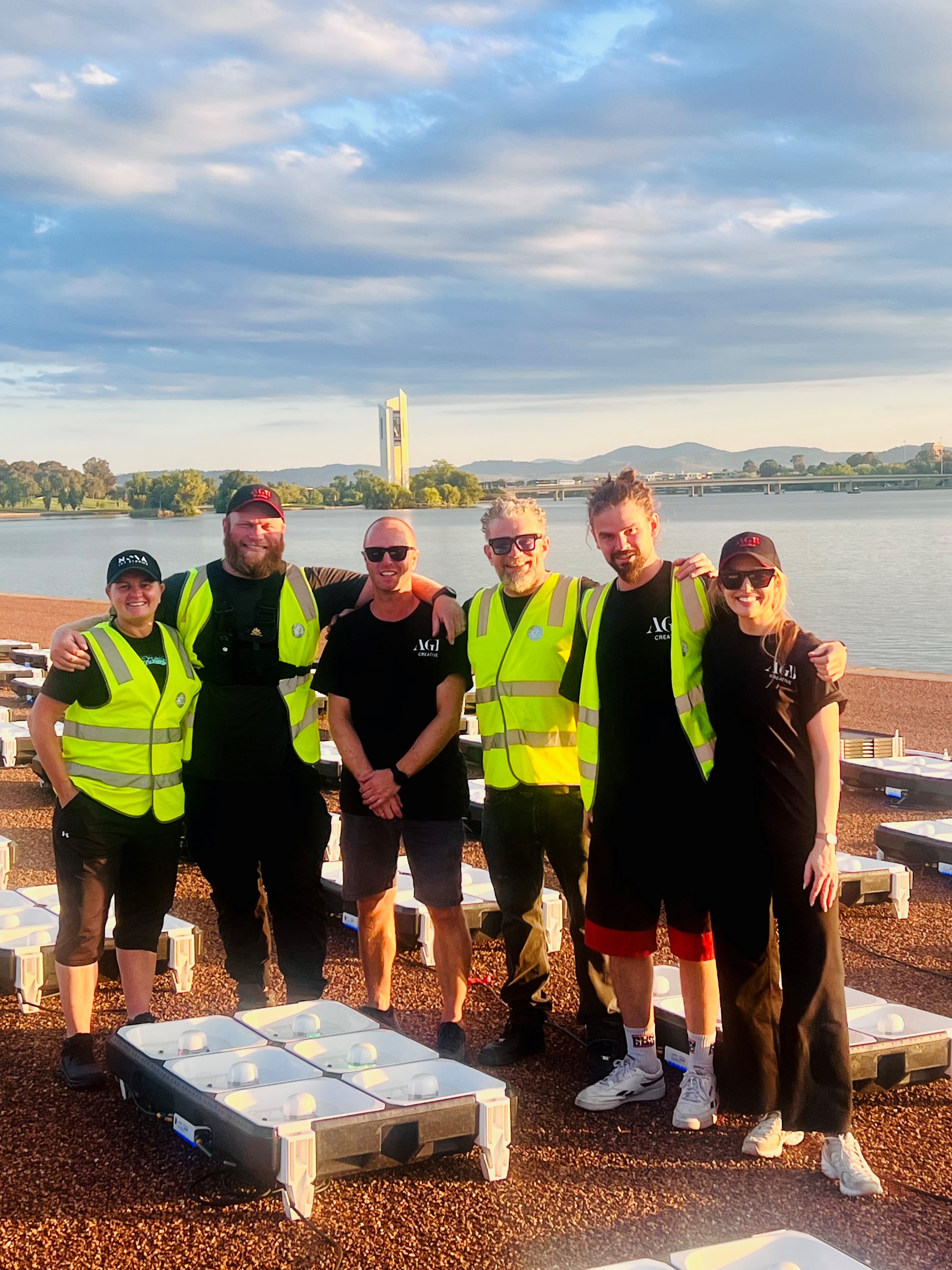 A group of people, some with high visibility vests, standing together in the middle of drones, docked on the ground with a lake, bridge and tower in the background.