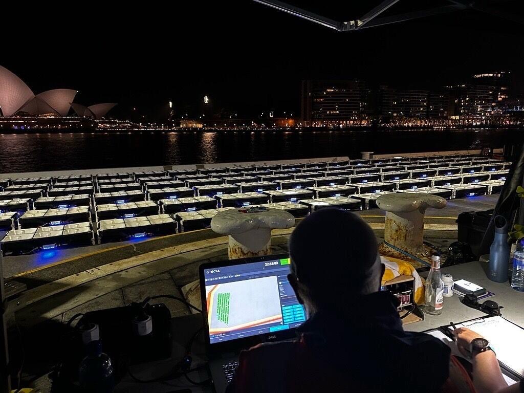 Alt text: A person stilling at a desk with a laptop in front of many drones docked on the ground with the Sydney Opera House in the background.